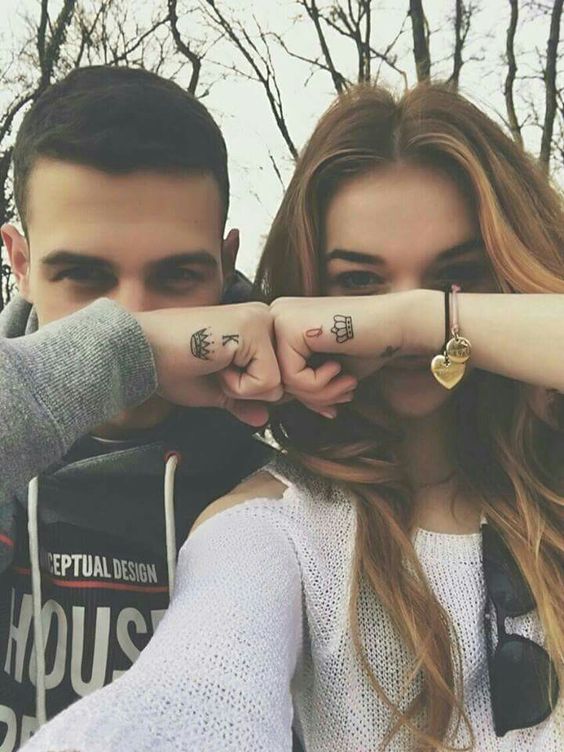 Casal exibindo tatuagens combinando nos pulsos com símbolos de rei e rainha. Fotos românticas ao ar livre.