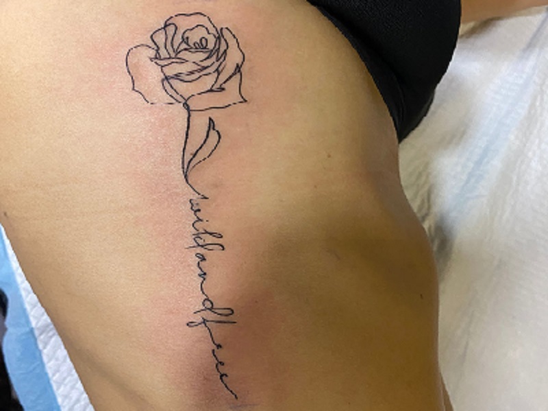 Tatuagem delicada de rosa com a frase wild and free no corpo.