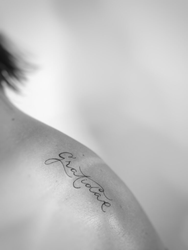 Tatuagem de caligrafia delicada no ombro com a palavra Gratidão em preto e branco.