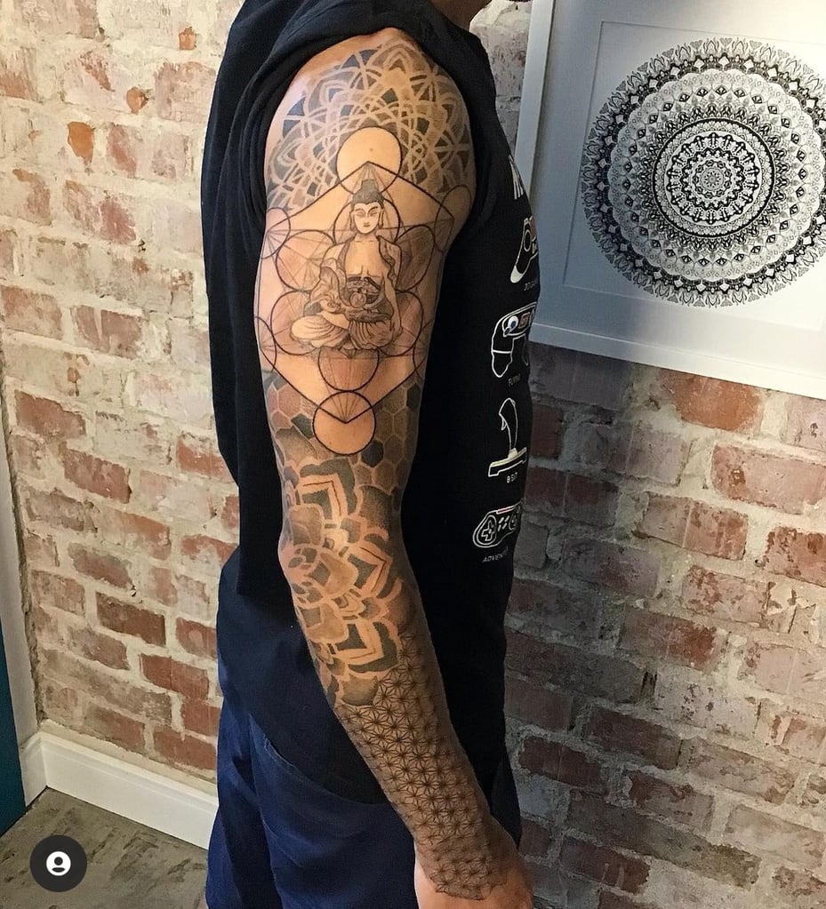 Homem com tatuagem de Buda e padrões geométricos em um braço, próximo a uma parede de tijolos e arte mandala.