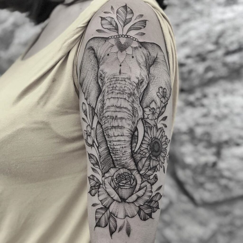 Tatuagem de elefante realista em preto e branco no braço, com flores e folhas detalhadas.