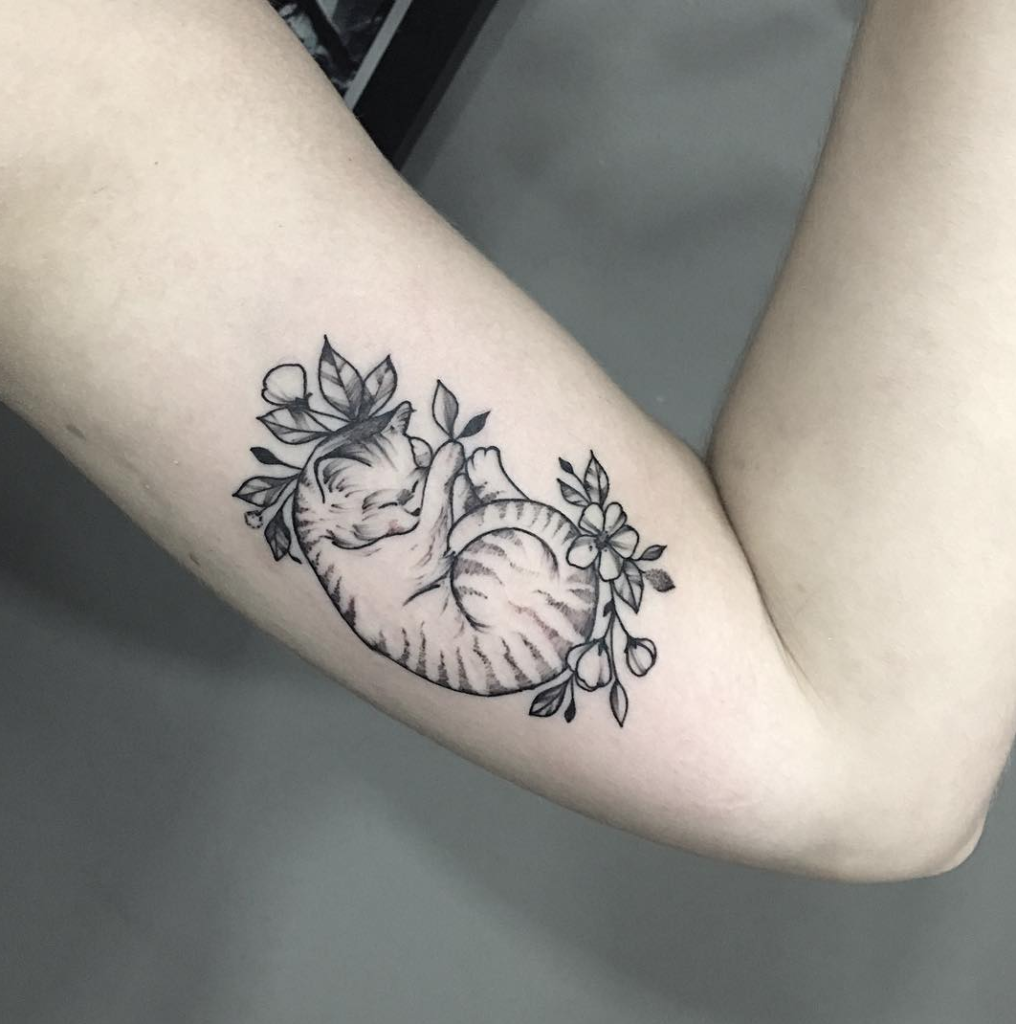 Tatuagem delicada de gato dormindo com flores no braço, em preto e cinza, estilo minimalista.