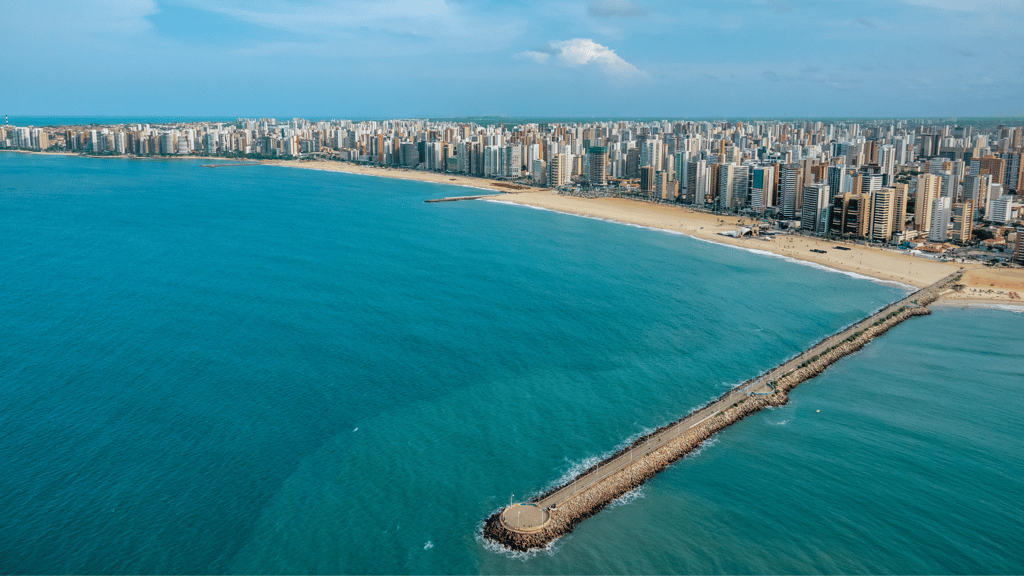 Imagem aérea de praia urbana, com mar azul e edifícios ao fundo, destacando um longo píer na cidade.
