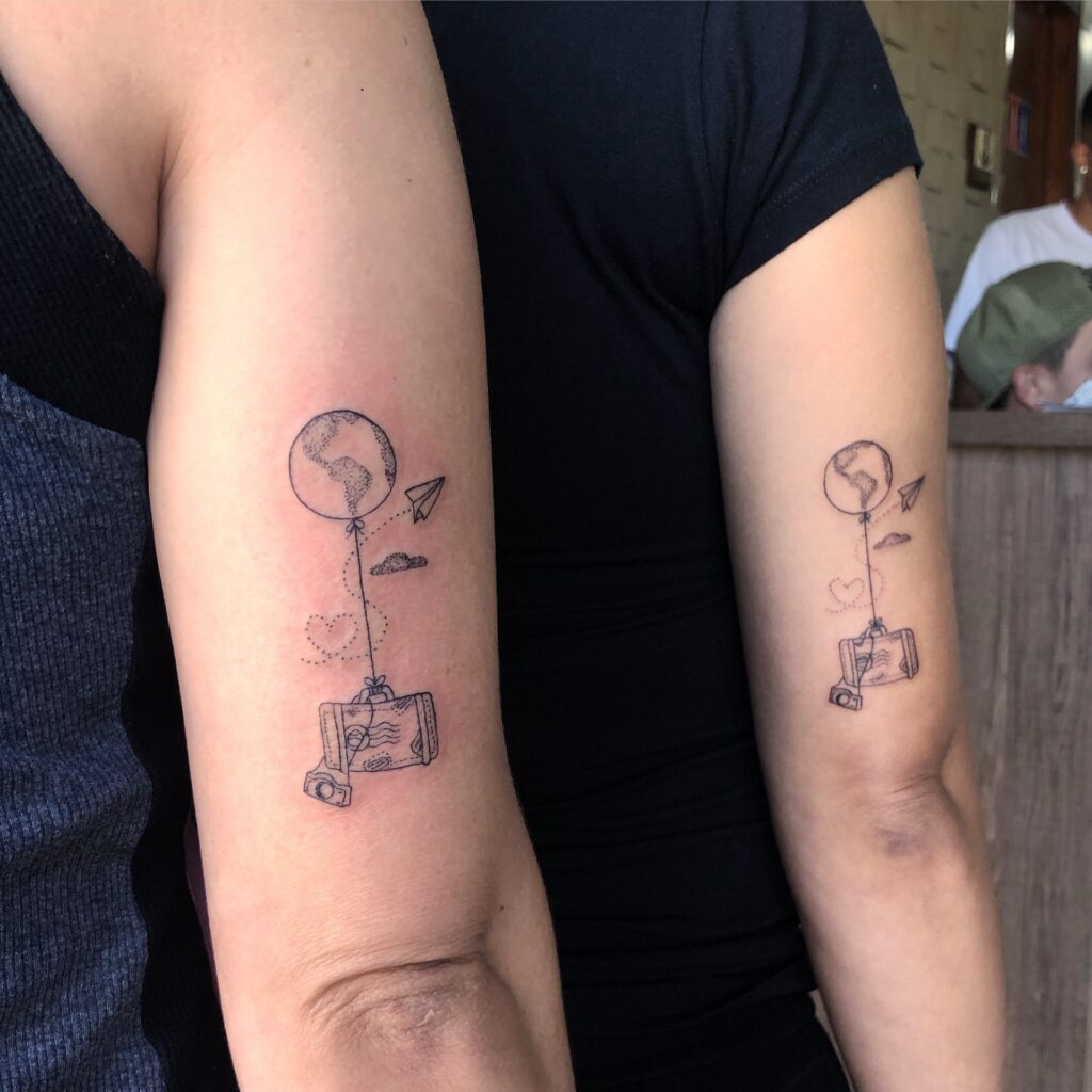 Tatuagem de viagem nas costas do braço com balão, mala e câmera. Símbolos de aventura e exploração.