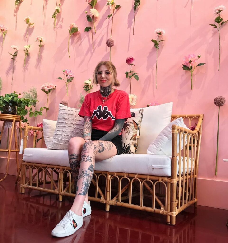 Mulher tatuada sentada em sofá de vime, parede rosa com flores atrás, camiseta vermelha e tênis branco.