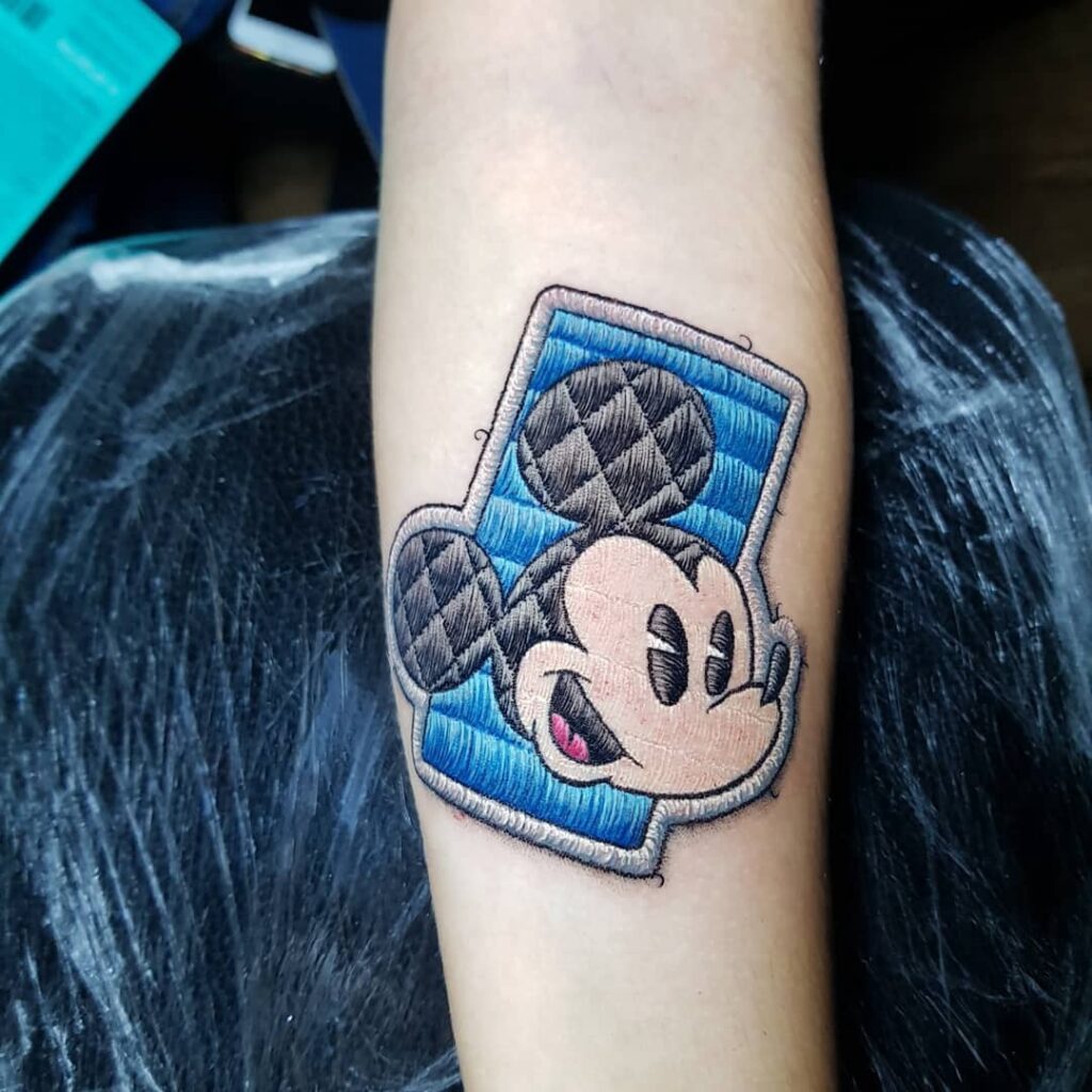 Tatuagem de personagem famoso estilo bordado no braço, com detalhes em tons de azul e preto.