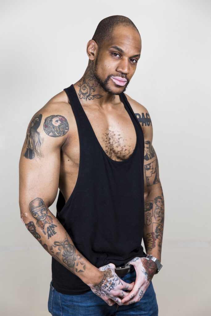 Homem tatuado com camiseta preta e calça jeans, com tatuagens nos braços e pescoço, posando para foto.