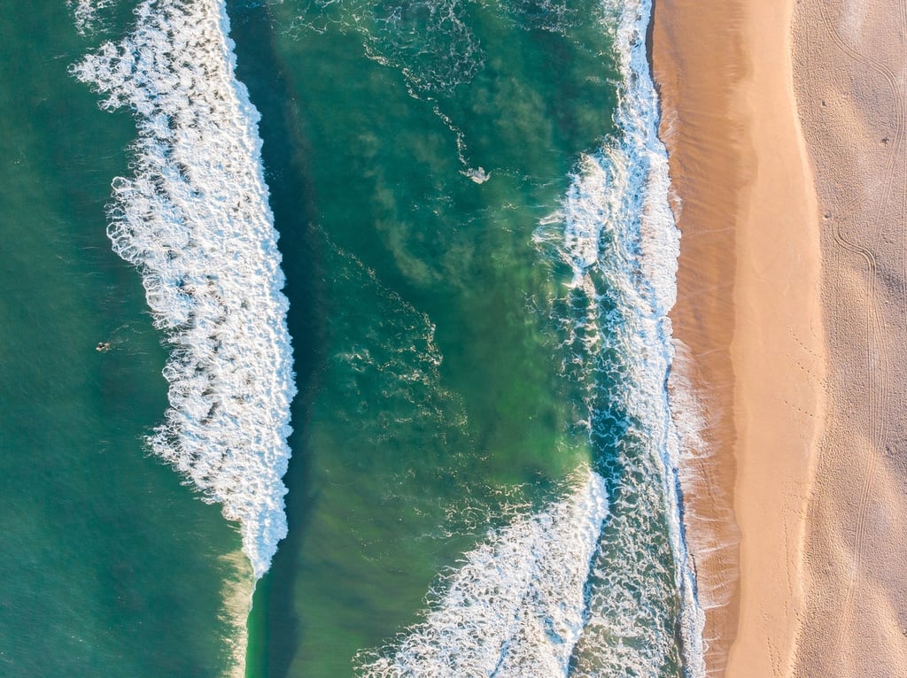 Vista aérea de ondas do mar azul e praia de areia dourada, destacando a beleza natural da costa.