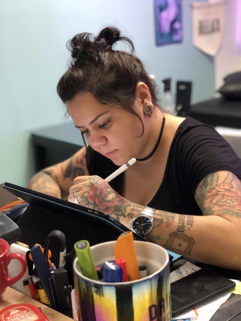 Pessoa concentrada desenhando em tablet, cercada por canetas e marcadores em escritório colorido.