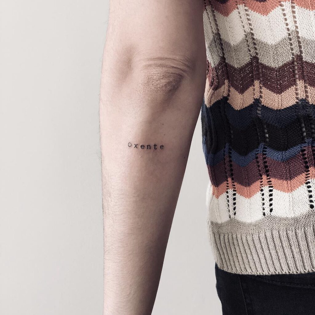 Braço com tatuagem escrita oxente em fonte simples, ao lado de blusa colorida de tricô com padrão geométrico.