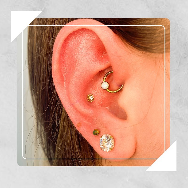 Close-up de orelha com piercings dourados e brilhantes, incluindo um brinco circular com pedra branca.