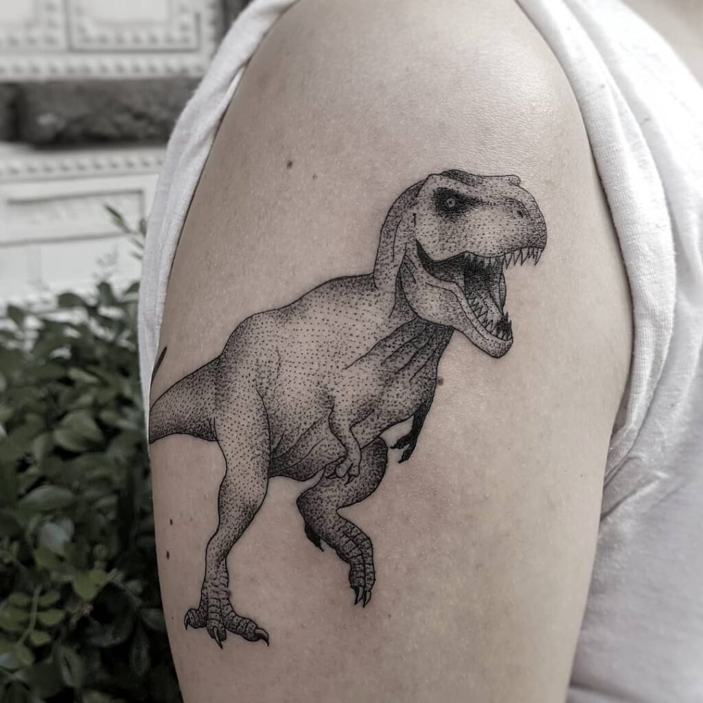 Tatuagem de Tiranossauro rex realista no braço de uma pessoa usando camiseta branca.