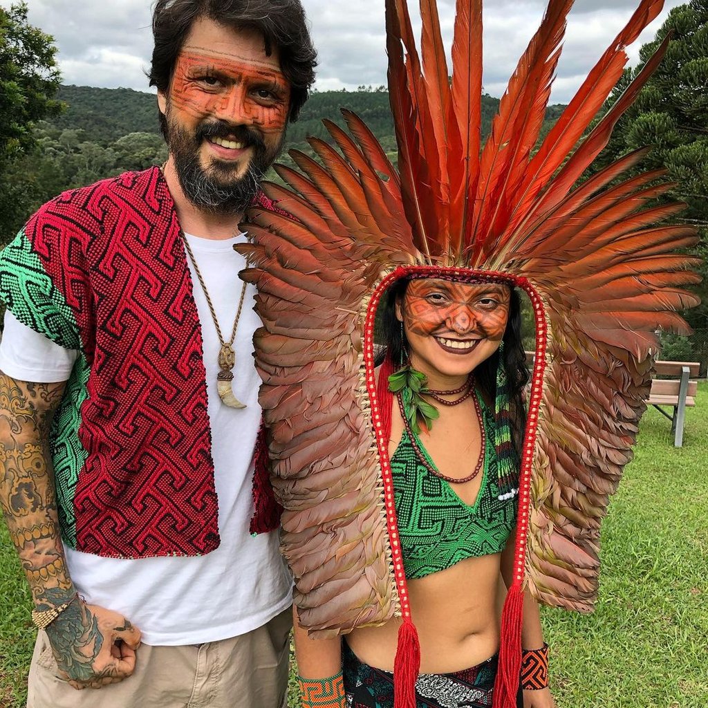 Duas pessoas sorrindo vestem trajes tradicionais com pintura corporal e cocares de penas em ambiente natural.