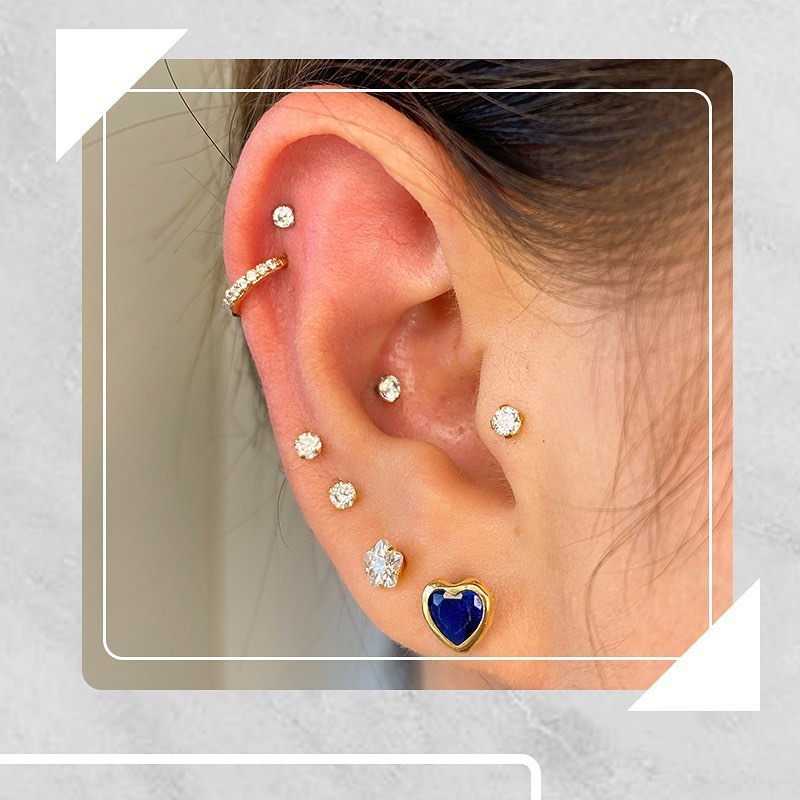Orelha com múltiplos piercings de diamante e brinco de coração azul dourado, destacando estilo e elegância.