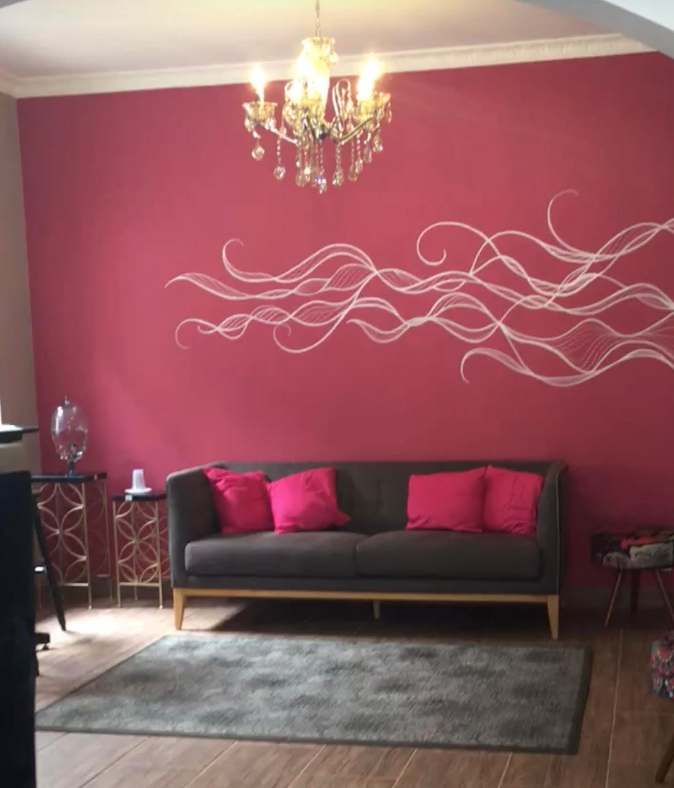 Sala elegante com sofá cinza, almofadas rosas, parede rosa decorada e lustre de cristal.