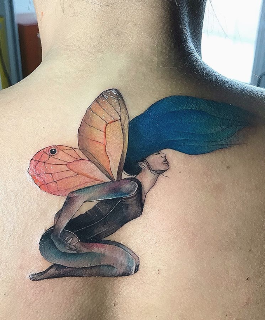 Tatuagem de fada com asas coloridas e cabelo azul em ombro feminino, representando fantasia e beleza.