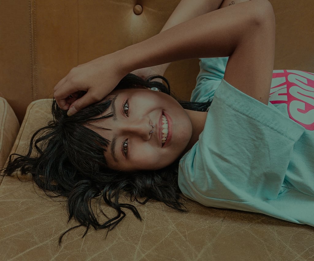 Pessoa deitada em sofá marrom, sorrindo e relaxando, usando camiseta azul clara.