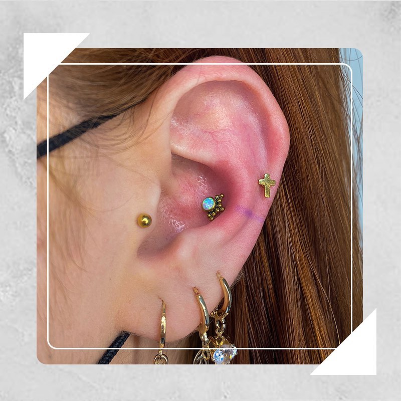 Orelha com diversos piercings dourados, incluindo cruz e pedras brilhantes. Estilo moderno e elegante.