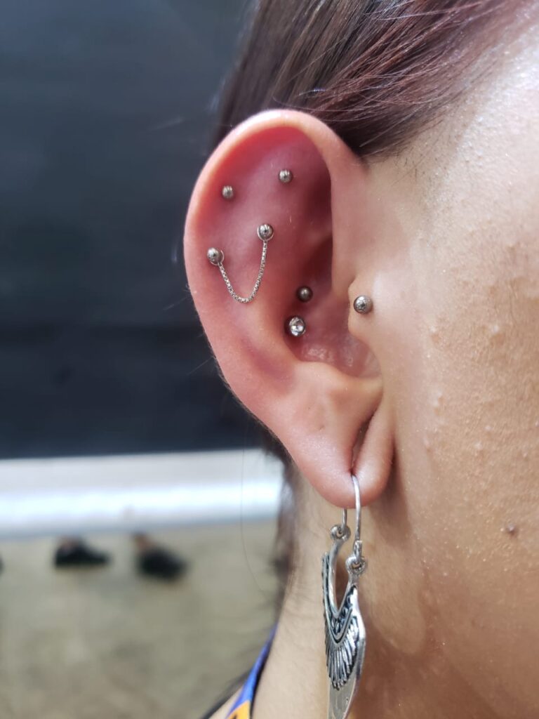 Orelha com múltiplos piercings prateados e corrente, estilo moderno de acessórios.