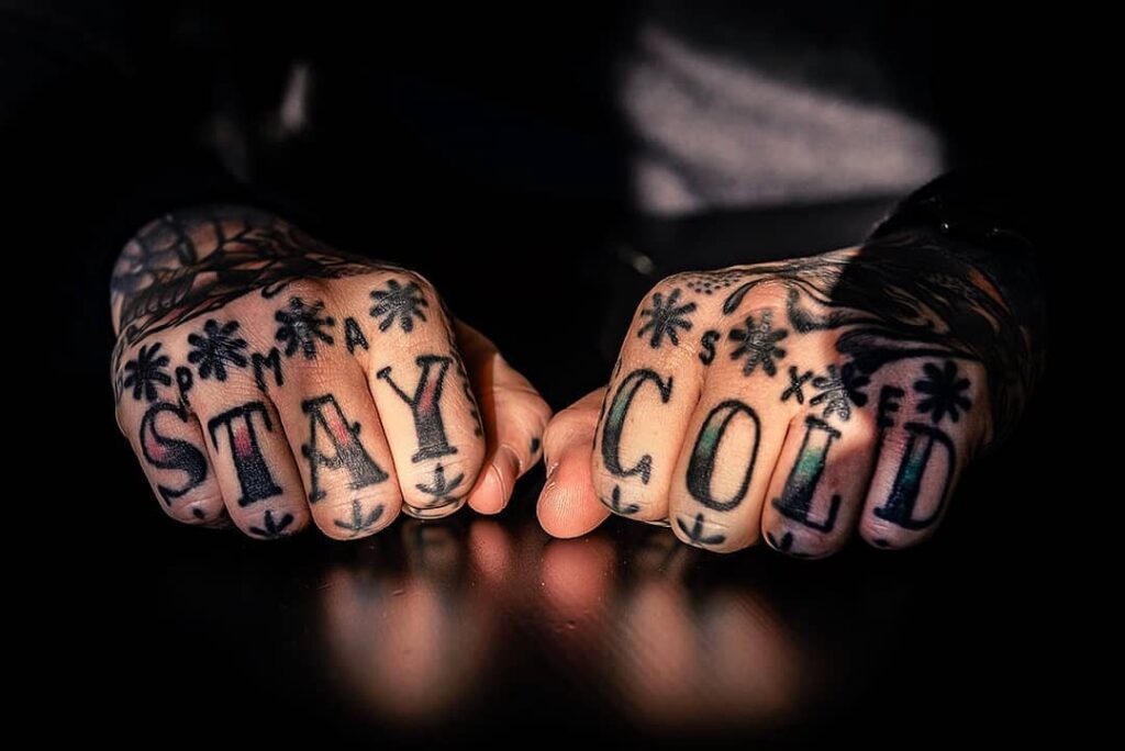 Punhos tatuados com as palavras Stay Cold em ambiente com iluminação dramática.