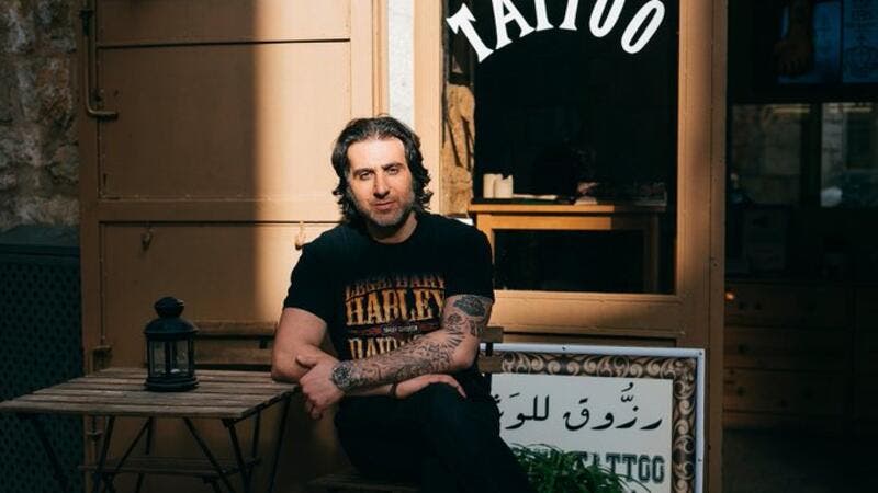 Homem sentado em frente a estúdio de tatuagem, exibindo tatuagens no braço e usando camiseta Harley-Davidson.