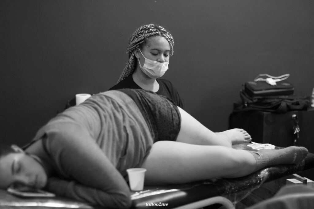 Pessoa recebendo tatuagem em estúdio, profissional concentrada com máscara, ambiente em preto e branco.