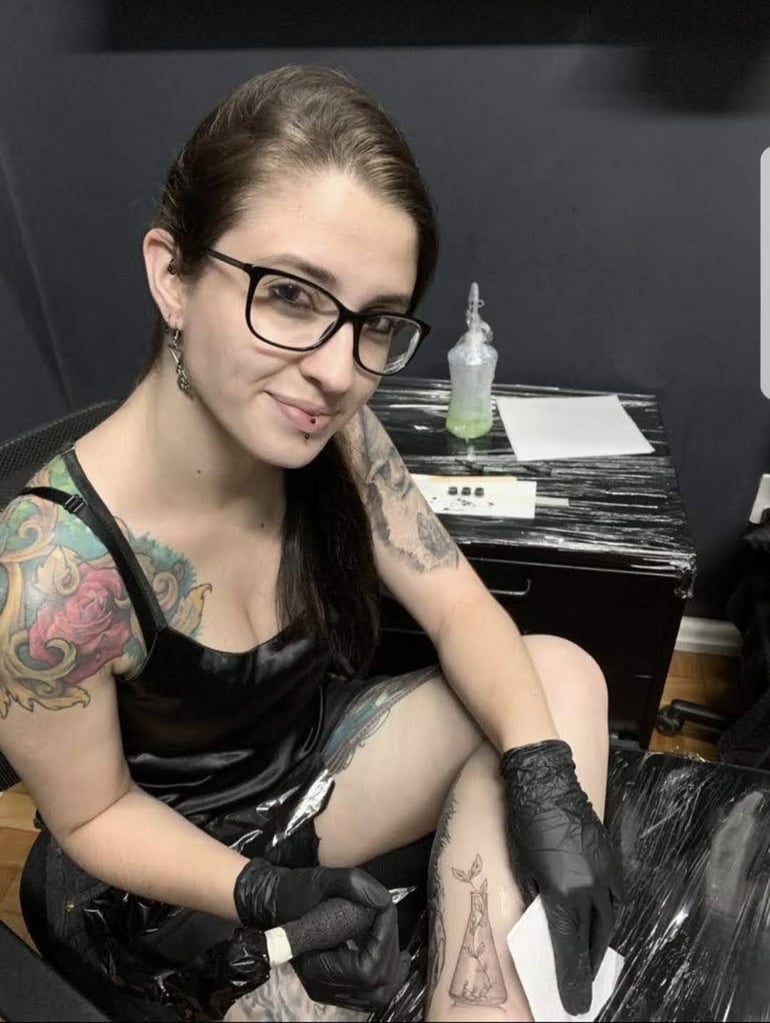 Artista com tatuagens coloridas, usando luvas, trabalha em design detalhado na perna em estúdio de tatuagem.