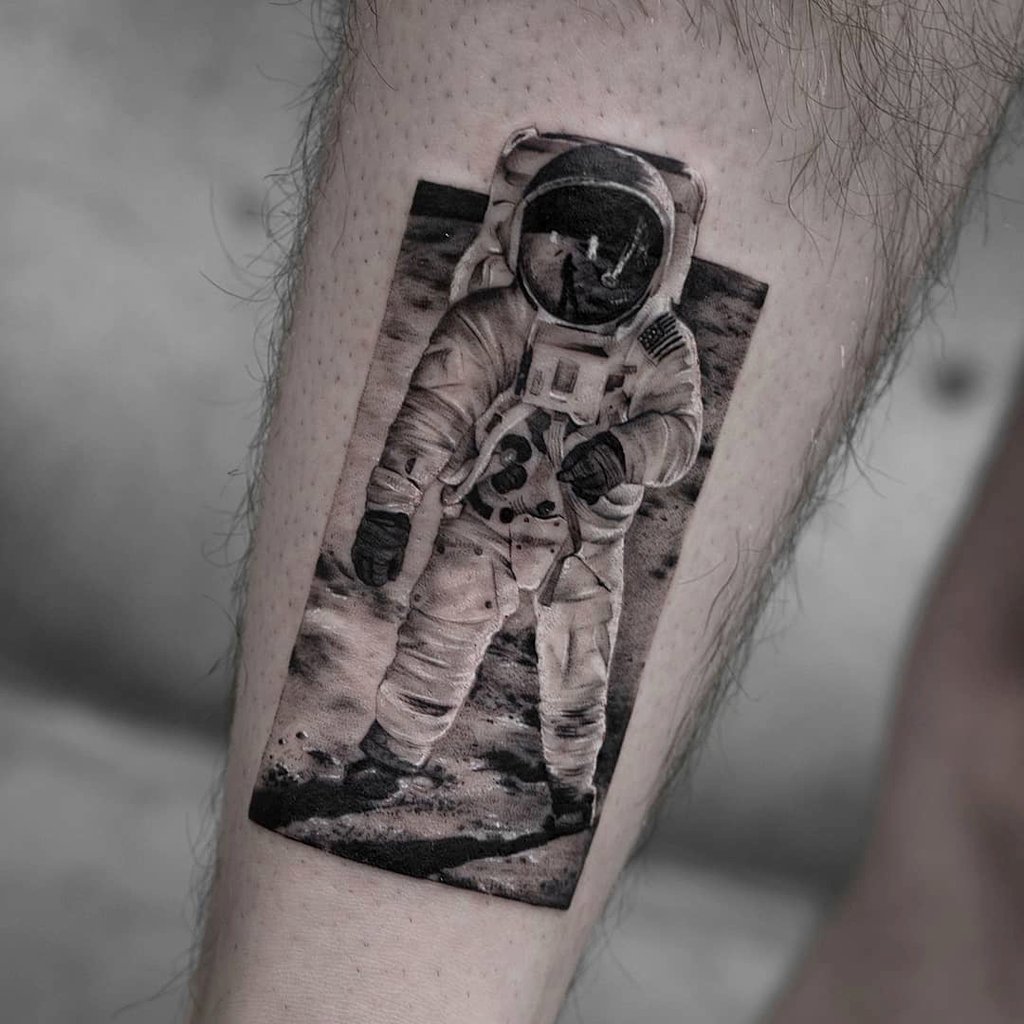 Tatuagem de astronauta em traje espacial na lua, retratada em preto e branco na perna de uma pessoa.