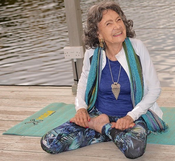 Mulher idosa praticando yoga em um deck à beira do rio, vestindo roupas coloridas e sorrindo.