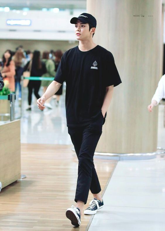 Homem caminhando em aeroporto vestindo roupa casual preta e boné. Ambiente moderno e movimentado ao fundo.