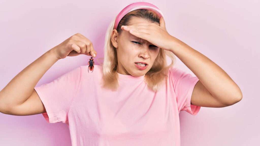 mulher branca e loira segurando uma barata com a ponta dos dedos da mão direita em fundo rosa