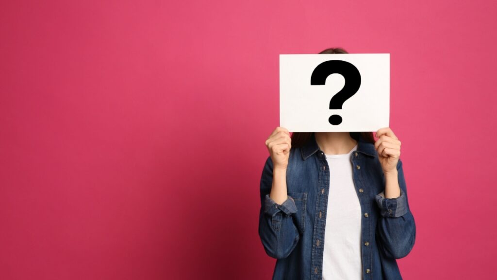 à direita está uma mulher segurando uma placa com um ponto de interrogação tampando a cara em fundo pink
