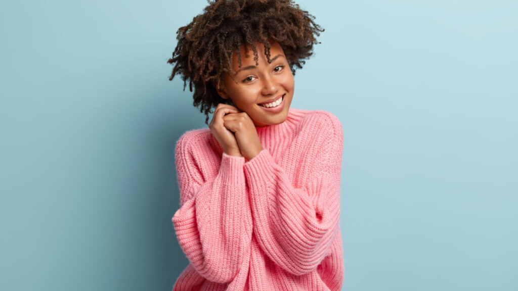 Retrato de uma mulher alegre, com uma expressão facial despreocupada, com um sorriso gentil, mantendo as mãos juntas perto do rosto, sentindo-se feliz.