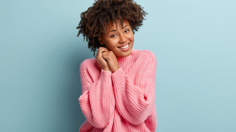 Retrato de uma mulher alegre, com uma expressão facial despreocupada, com um sorriso gentil, mantendo as mãos juntas perto do rosto, sentindo-se feliz.