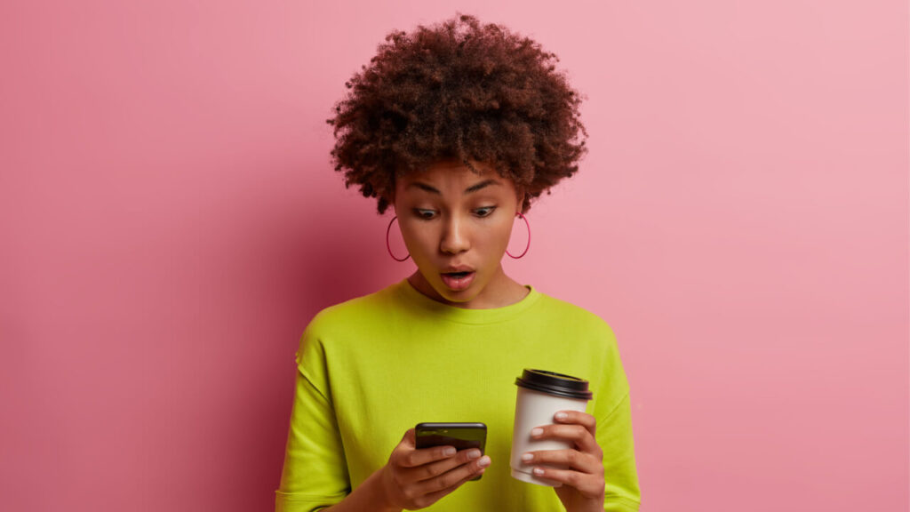 Mulher de cabelos cacheados surpresa, mexendo no celular, lendo algo chocante ou inacreditável, expressando espanto, focada no celular, segurando um copo de café para viagem, isolada em uma parede rosa.