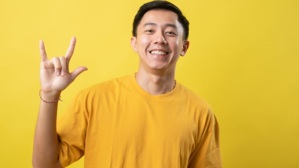 homem jovem com traços asiáticos fazendo um sinal de eu te amo em libra em fundo amarelo