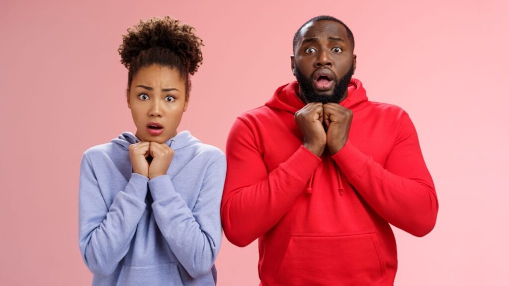 Uma mulher e um homem preocupados, encolhidos, pressionando as mãos no peito, com os olhos arregalados, em pé, nervosos, em um fundo rosa. Conceito de emoções intensas.