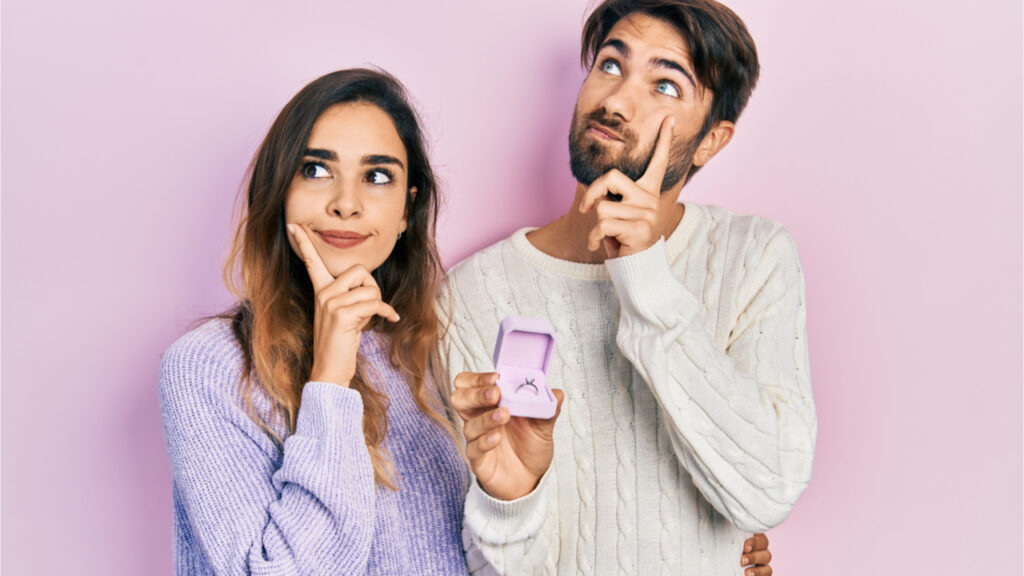 Casal de namorado e namorada segurando o anel de noivado para proposta. Cara séria, pensando em questão com a mão no queixo, pensativos sobre a ideia confusa.