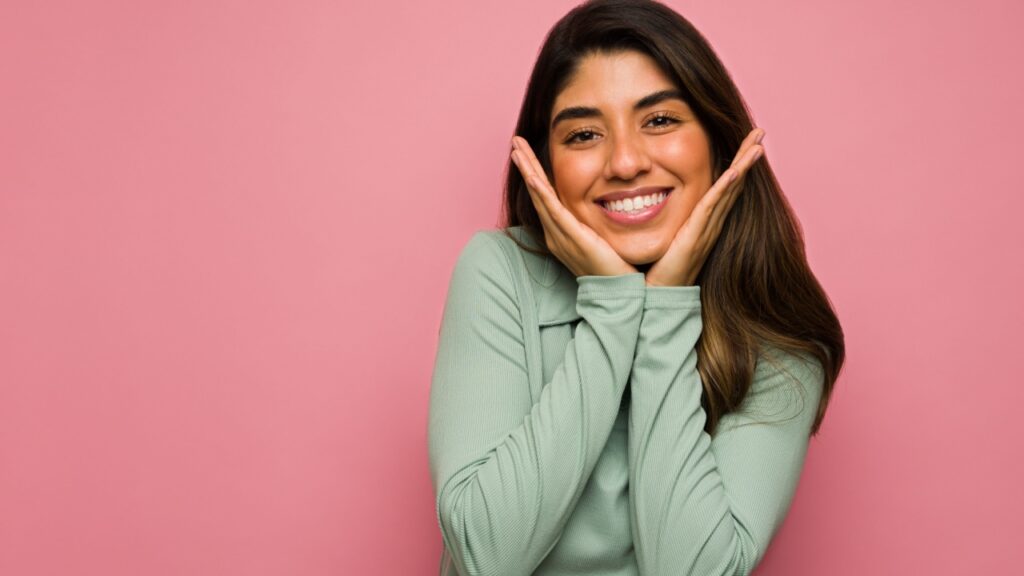 mulher sorrindo com a mão no queixo em fundo rosa