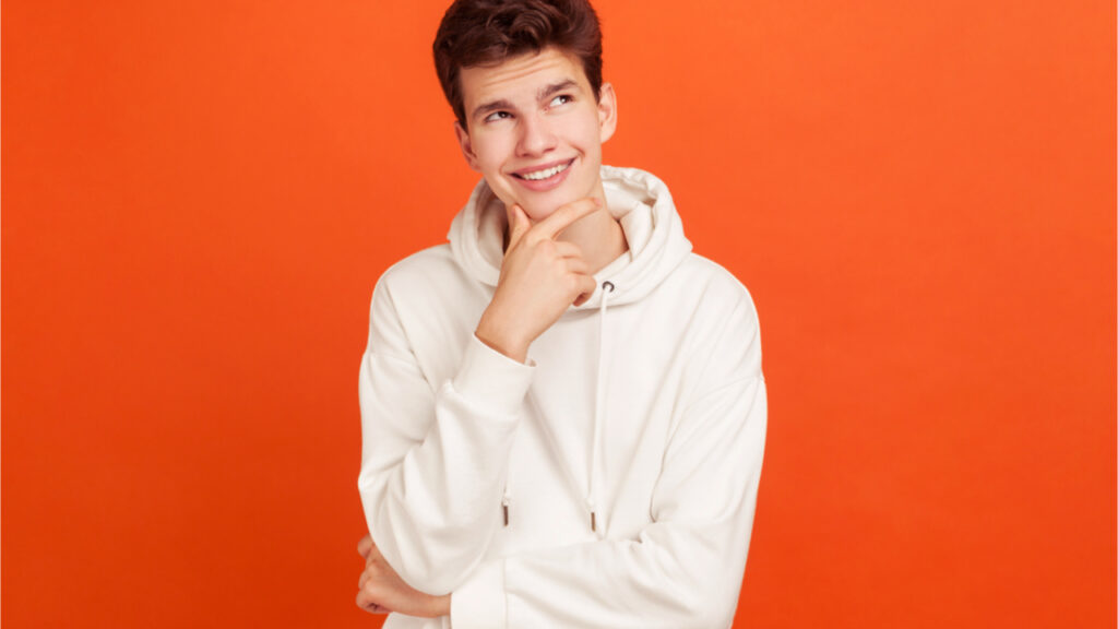 Jovem sorridente com rosto pacífico pensando em planos para o futuro segurando seu queixo. Adolescente pensativo. Foto de estúdio no interior isolado em fundo laranja.