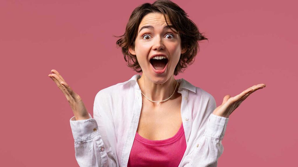 mulher com expressão facial de animada com os braços na altura dos ombros e boca aberta em fundo rosa