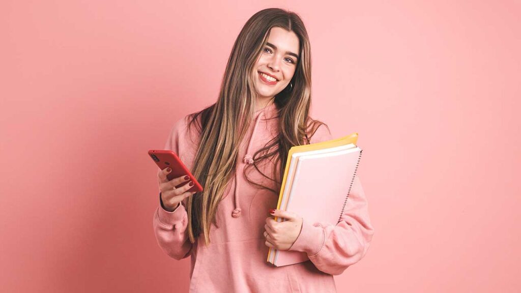 mulher jovem segurando duas apostilas no braço esquerdo e uma celular na mão direita usando moletom rosa em fundo rosa