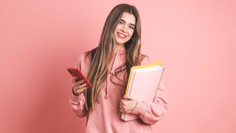 mulher jovem segurando duas apostilas no braço esquerdo e uma celular na mão direita usando moletom rosa em fundo rosa