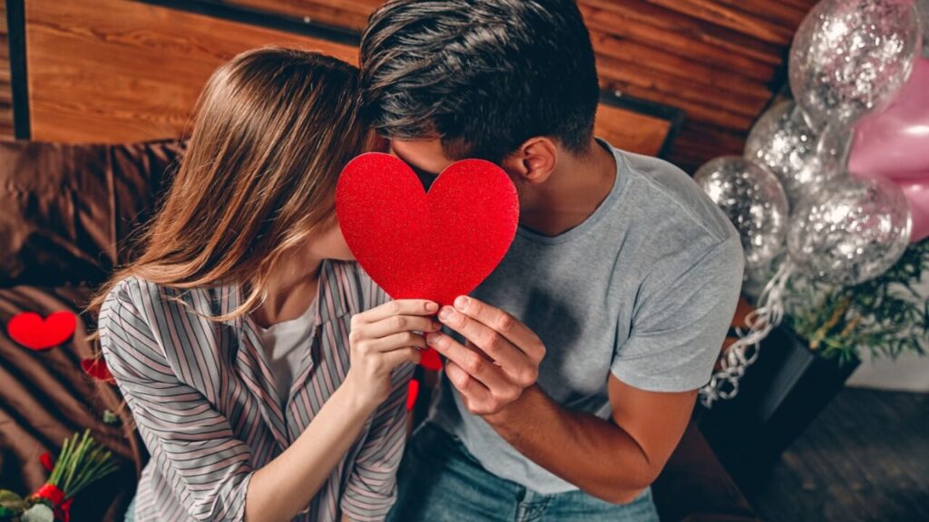 casal heterossexual sentados no sofá com um coração vermelho cobrindo os rostos que estão juntos