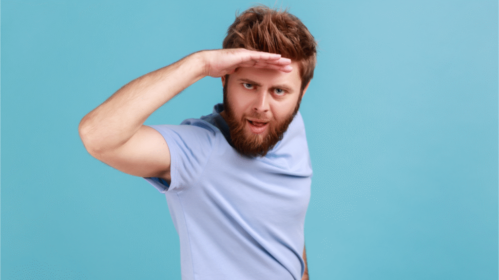 Retrato de homem barbudo adulto jovem bonito olhando longe com a mão sobre a cabeça, vendo algo agradável. Foto de estúdio interior isolada sobre fundo azul.