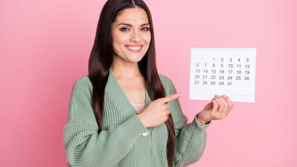 Foto de mulher segurando um calendário e apontando para ele com o dedo direto, usando um casaco de lã verde, isolada em um fundo de cor rosa pastel.