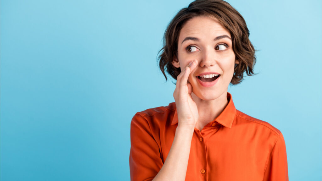Foto de mulher segurando o braço perto da boca, olhando para o lado, contando um segredo, usando camisa laranja, isolada no fundo de cor azul.