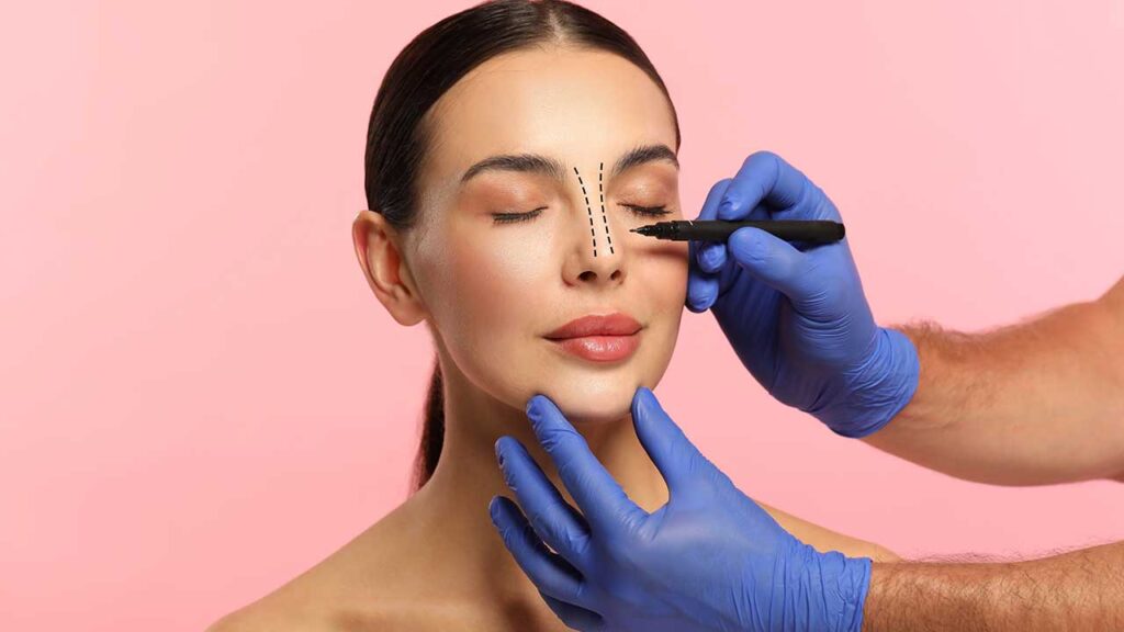 par de mãos com luva azul fazendo marcação com caneta preta no nariz de uma mulher jovem em fundo rosa