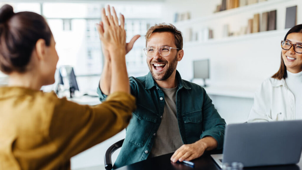 homem de óculos fazendo high five em seu colega de escritório em um ambiente branco com alguns livros