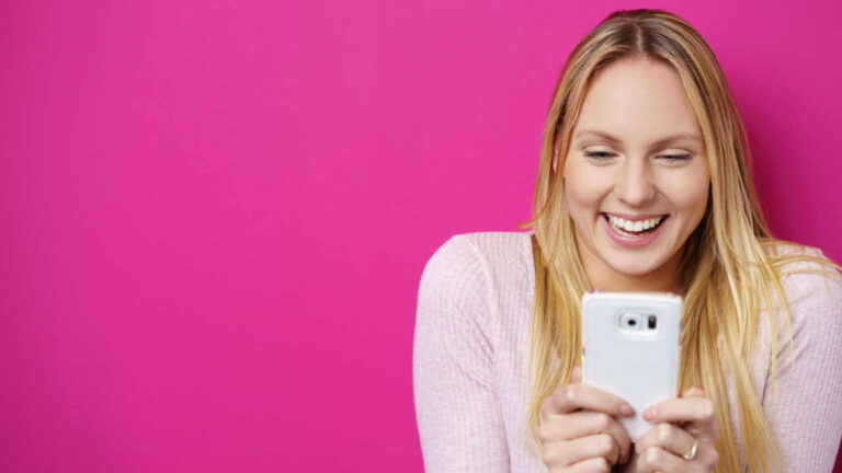 Mulher loira feliz olhando para seu celular com um sorriso surpreso contra um fundo rosa no estúdio.